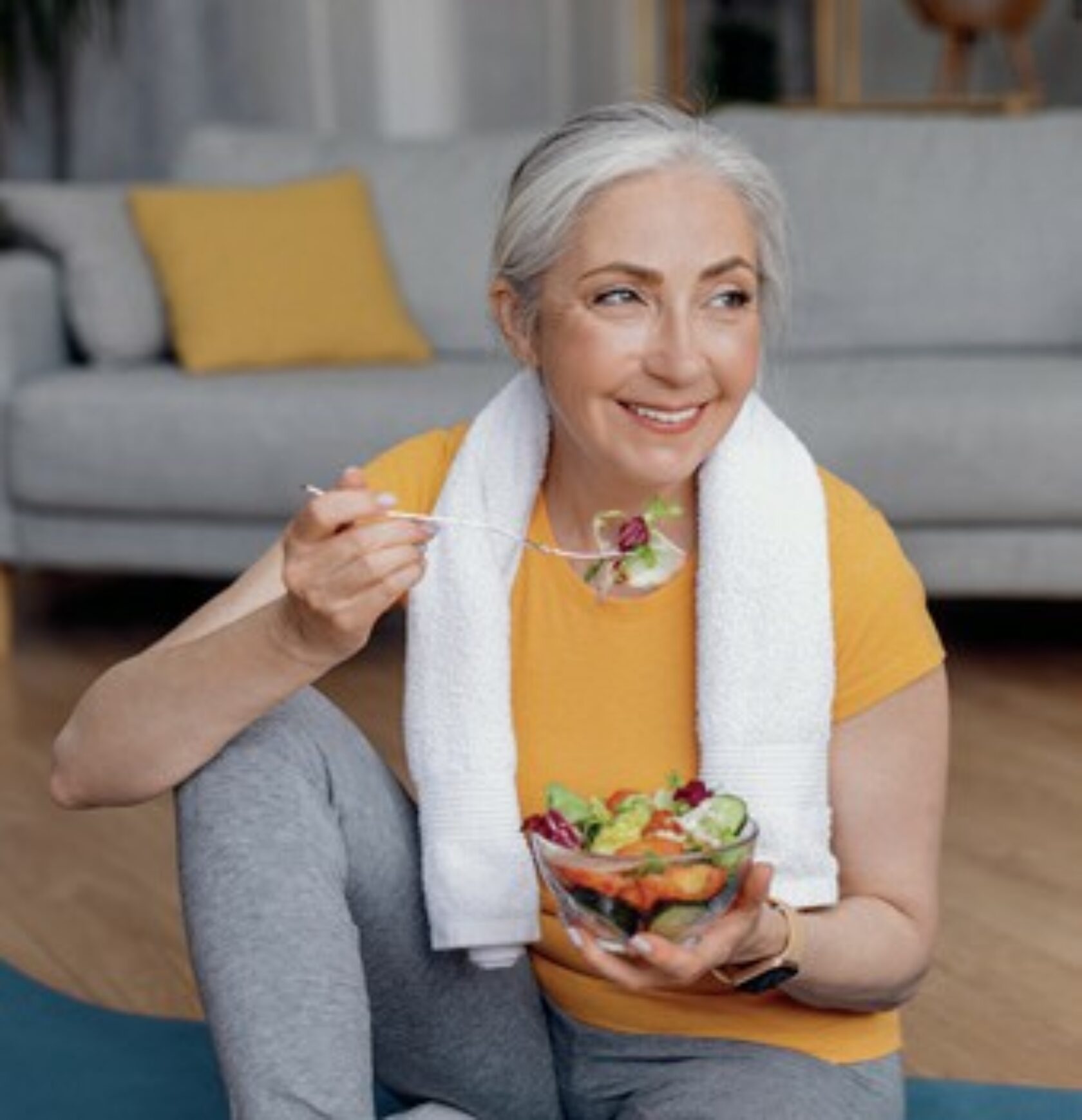Grauhaarige Frau isst nach dem Training einen Salat. Sie hat noch ein Handtuch um die Schultern und Sportkleidung an.