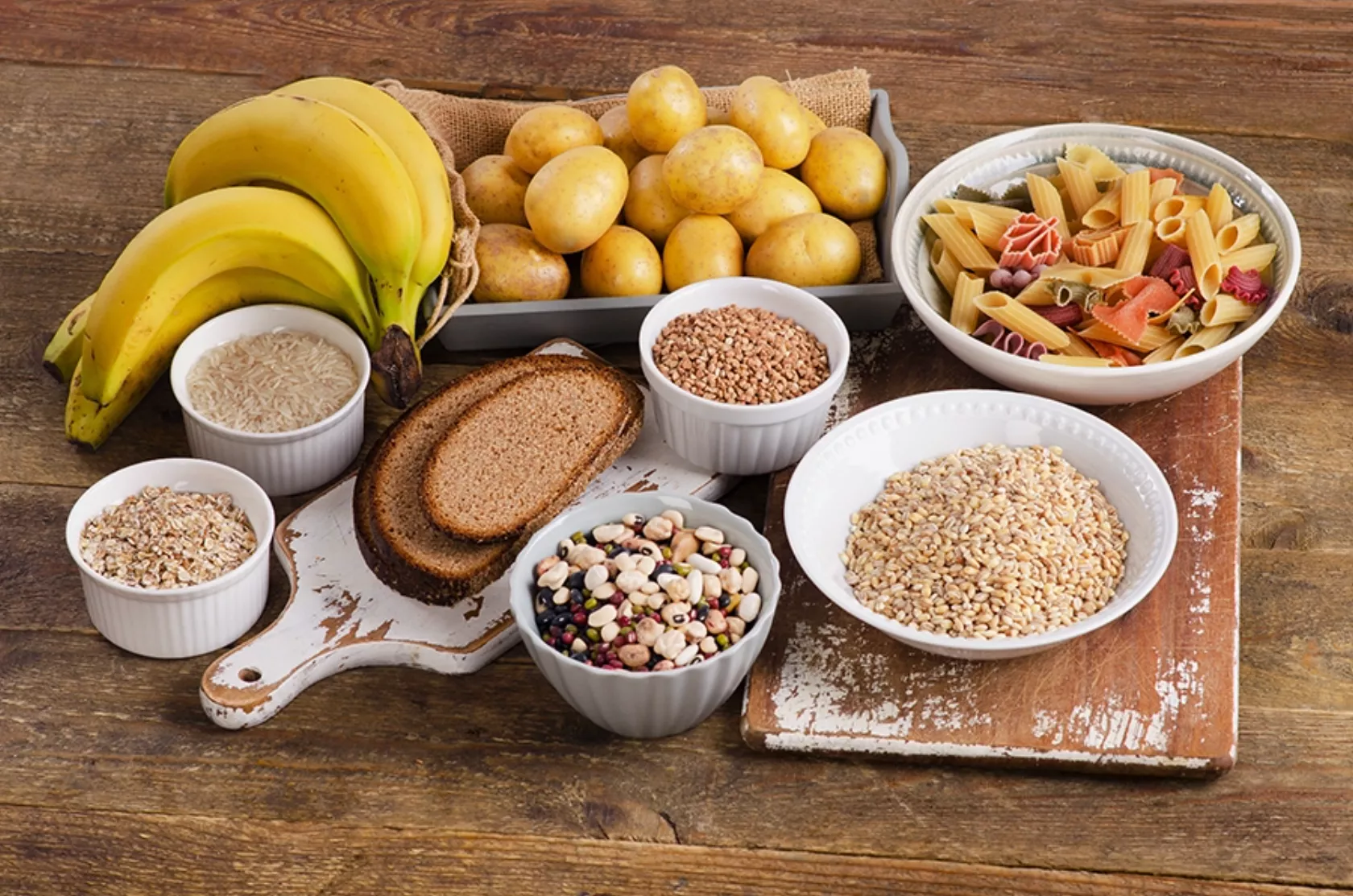 Kohlenhydratreiche Nahrungsmittel wie Bananen, Kartoffeln, Brot, Getreide und Hülsenfrüchte auf einem Tisch angeordnet.