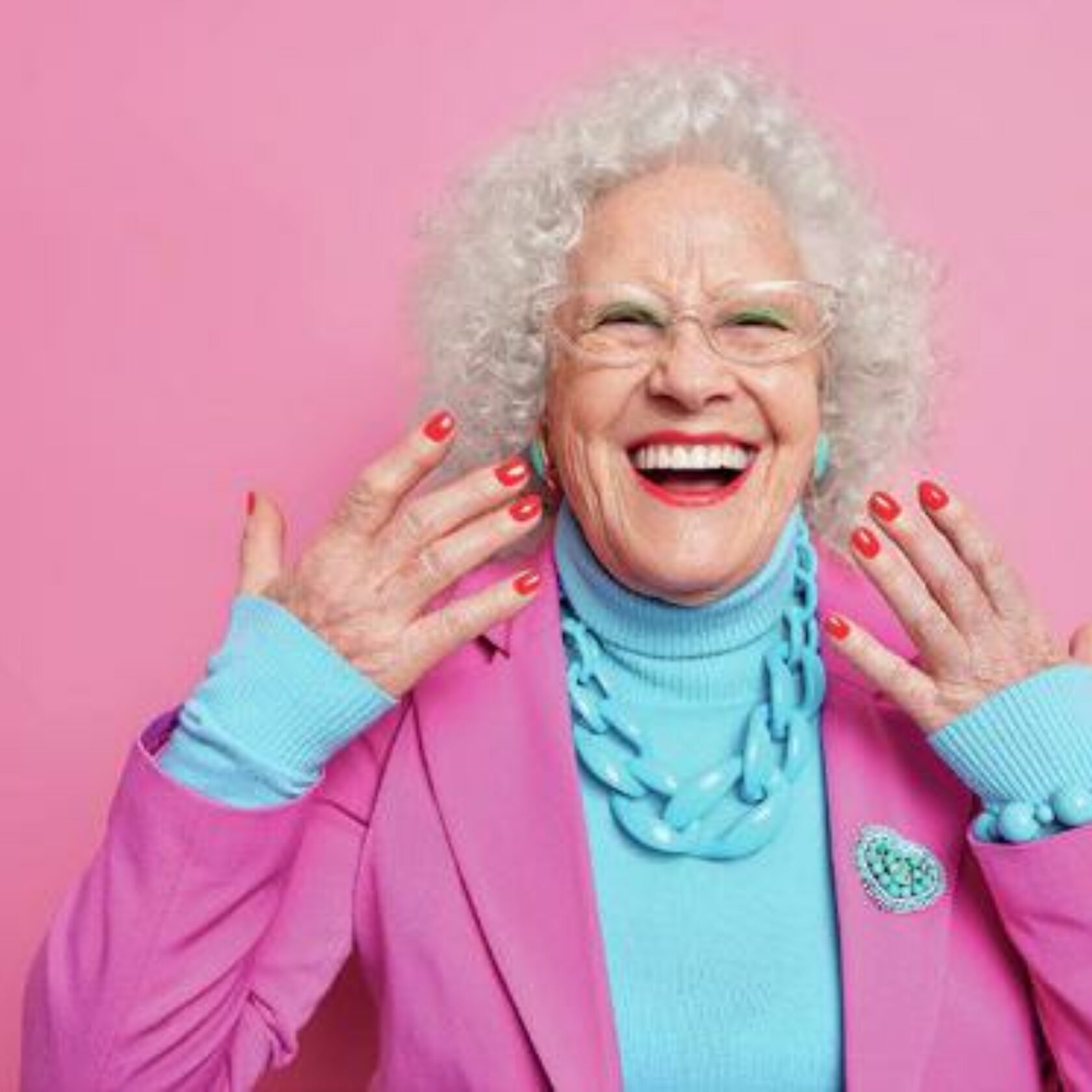 Eine Seniorin mit verhältnismässig junger Haut und rot lackierten FIngernägeln und pink-blauem Outfit lacht in die Kamera.