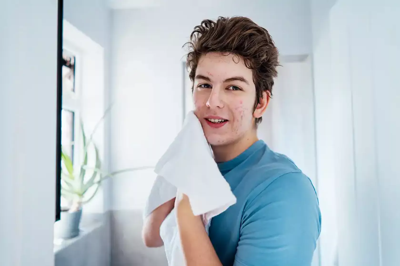 Junger Mann mit Akne im Gesicht trocknet sich nach der Reinigung das Gesicht mit einem Handtuch und blickt in die Kamera..