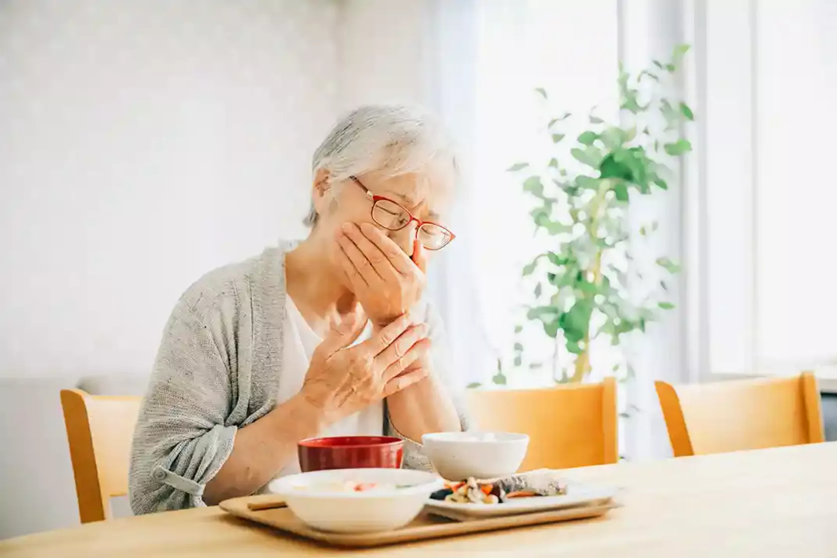 Seniorin sitzt vor ihrem Essen und hält die Hand vor den Mund.