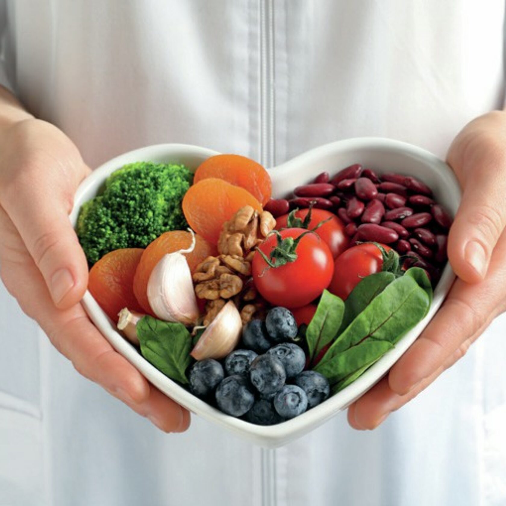 Lebensmittel, die gesund fürs Herz sind - wie Brokkoli, Aprikosen, Knoblauch, Walnüsse, Bohnen, Tomaten, Heidelbeeren - in einer herzförmigen Schale