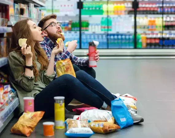 Zwei junge Erwachsene sitzen im Supermarkt auf dem Boden und stopfen sich hemmungslos Chips und andere hochverarbeitete Lebensmittel in den Mund.