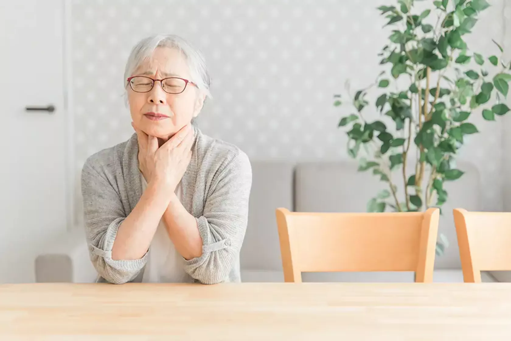 Eine Seniorin sitzt am Tisch und hält sich mit beiden Händen den schmerzenden Hals.